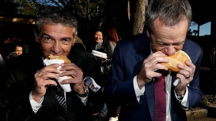 Opposition Leader Bill Shorten eats a sausage in a roll with onion and tomato sauce, together with ALP candidate for Reid, Angelo Tsirekas, during a visit to the polling booth at Strathfield North Public School in the seat of Reid, on Saturday 2 July 2016. Photo: Alex Ellinghausen